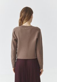 Brauner, gestrickter Pullover mit Rundhalsausschnitt, gerippten Schulterakzenten und einem subtilen Knopfdetail auf der Rückseite. Weicher Stoff und taillierte Passform.