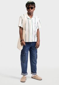 Kurzärmliges gestreiftes Hemd in Creme, Grün und Marineblau, kombiniert mit blauen Cargo-Jeans, beigen Clogs und einer grünen Mütze. Trägt eine Canvas-Tote-Tasche.