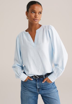 Femme avec les mains dans les poches portant une blouse à manches longues de couleur bleu clair avec un col en V et un jean bleu, sur un fond uni.