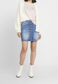 Stickad krämfärgad kofta, ljusrosa topp med silvergrafik, blå denim skjortkjol, vita cowboystövlar med präglad textur.