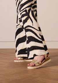 Gouden metallic sandalen met een brede band, witte zool en gestructureerde details. Gecombineerd met zwart-witte zebra-patroon broeken.