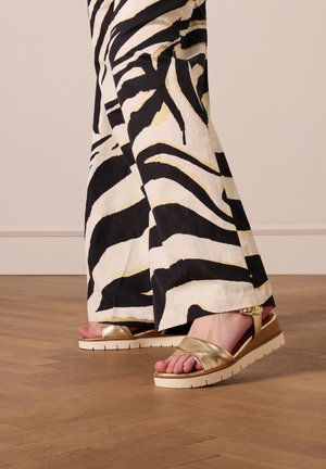 Sandales métalliques dorées avec une large bride, semelle blanche et détails texturés. Assorties à un pantalon à motif zèbre noir et blanc.