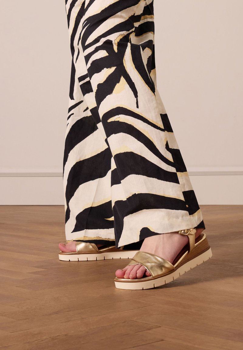 Gouden metallic sandalen met een brede band, witte zool en gestructureerde details. Gecombineerd met zwart-witte zebra-patroon broeken.