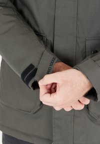 Giacca impermeabile grigia con polsini in velcro e etichetta ricamata. Presenta una chiusura frontale con bottoni e tasche laterali, mostrando una texture di tessuto liscia.