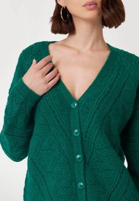Cardigan verde lavorato a maglia con una profonda scollatura a V, motivi testurizzati e cinque bottoni turchesi sul davanti. Tessuto morbido e caldo.