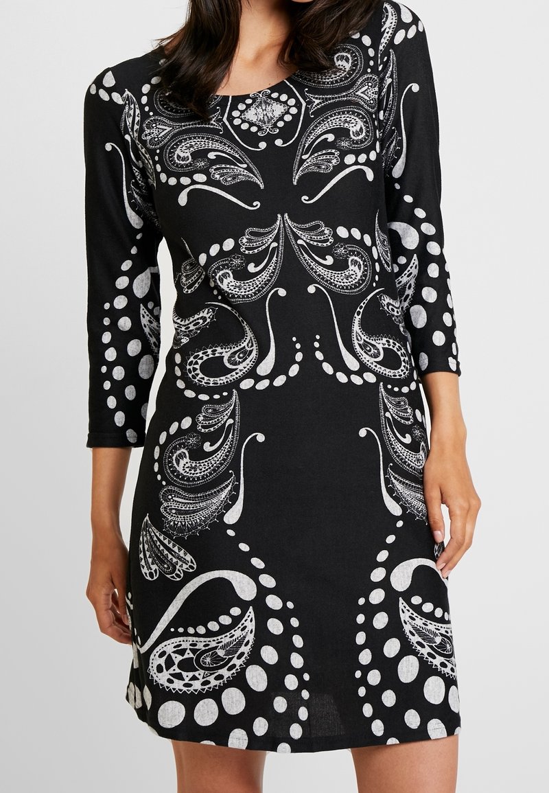 Femme portant une robe noire avec des motifs paisley et à pois blancs complexes, manches trois-quarts, visible des épaules jusqu'au milieu de la cuisse.