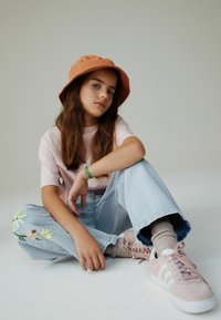 Maglietta di cotone rosa, jeans azzurri chiaro ricamati con motivi floreali, cappello arancione stile secchiello e sneakers rosa con strisce bianche.