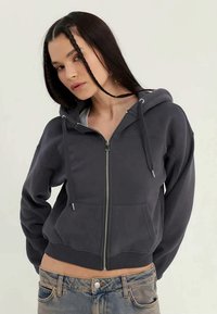 Tmavě šedá mikina na zip s přední kapsou, kapucí se stažením, ribbovanými manžetami a mírně zkráceným střihem. Složení z bavlny.