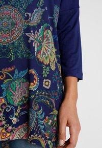 Tunique à motifs floraux multicolores sur fond marine, avec des manches trois-quarts et un tissu doux et texturé.