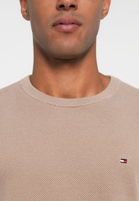 Prim-plan al unei persoane care poartă un pulover tricotat texturat bej, cu un mic logo roșu, alb și bleumarin pe piept.