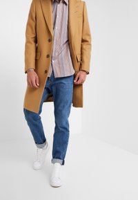 Mand iført kamel-frakke, stribet skjorte med knapper, opsmøgede blå jeans og hvide sneakers, går i en minimal hvid rum.