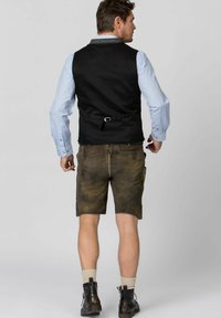 Schwarze Weste über einem hellblauen karierten Hemd, kombiniert mit braunen Lederhosen und dunklen Stiefeln. Die Weste hat einen Gürtel-Detail.
