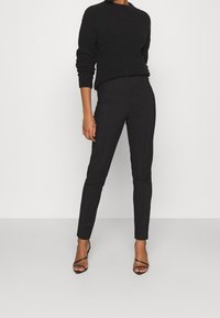 Czarny sweter o fakturowanej powierzchni i wysokim dekolcie, zestawiony z dopasowanymi czarnymi spodniami o eleganckim kroju i widocznych szwach.
