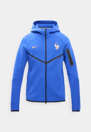 Modrá bunda s kapucňou a celým zipsom, čierne akcenty, logo Nike na pravej strane hrudníka a emblém Francúzskej futbalovej federácie na ľavej strane hrudníka.