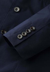 Rukáv námořnicky modrého obleku se čtyřmi tmavými knoflíky označenými "Hackett London", uspořádanými v řadě u manžety.