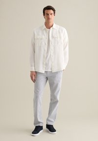 Camicia bianca in lino con bottoni, due tasche sul petto, abbinata a pantaloni a righe blu e bianche e sneakers scure, in piedi su uno sfondo neutro.