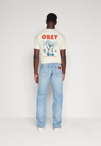 Tricou crem cu text și design grafic roșu "OBEY" pe spate, asortat cu blugi albaștri deschiși. Material din bumbac. Croi lejer.