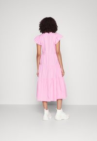 Różowa, warstwowa sukienka z falbanastymi rękawami, wykonana z lekkiego materiału. Modelka nosi białe tenisówki, a tło jest jednolite i szare.