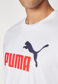 Valkoinen puuvillapaita, jossa on suuri punainen "PUMA"-logo ja musta hyppivä puma -grafiikka. Pyöreä kaula-aukko, lyhyet hihat, sileä pinta.