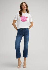 Biała koszulka z różowym okrągłym logo, zestawiona z przyciętymi, prostymi ciemnoniebieskimi dżinsami z postrzępionymi nogawkami i tylnymi kieszeniami.