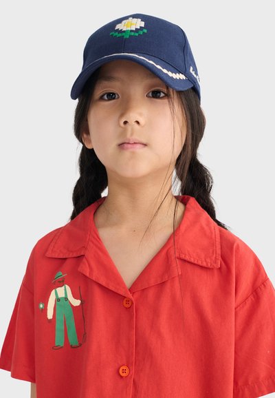Jeune fille avec deux tresses portant une casquette bleu marine ornée d'une fleur blanche et un t-shirt rouge avec l'image d'un fermier tenant une fourche.