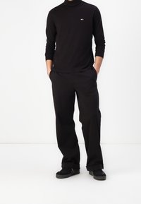 Black long-sleeve turtleneck, relaxed-fit black trousers, paired with black sneakers. Minimal logo detail on shirt, smooth fabric texture.