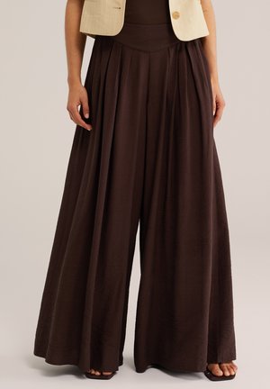 Mujer con pantalones plisados de pierna ancha y cintura alta de color marrón oscuro, combinados con un blazer corto de color beige claro.