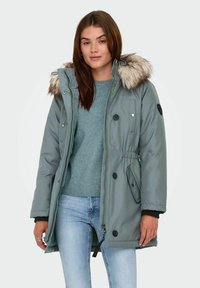 Parka grigia con bordo in pelliccia sintetica sul cappuccio, dotata di chiusura con zip e bottoni, tasche laterali e vita elasticizzata.