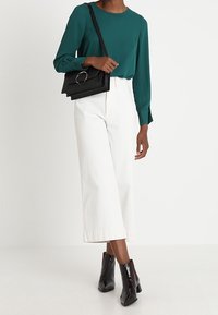 Blusa verde azulado con mangas largas, pantalones blancos de pierna ancha y bolso estructurado negro con un detalle circular. Botines negros brillantes.