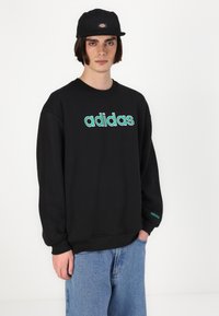 Sweatshirt preto com logótipo "adidas" verde, corte oversized, decote redondo, textura macia, combinado com jeans de ganga azul e um boné preto.