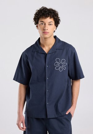 Jonge man met krullend haar die een marineblauw kortemouw overhemd draagt met een wit abstract bloemmotief op de borst.