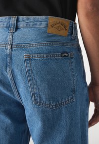 Blå denimjeans med rak passform, med en brun läderskylt, orange sömmar och två bakfickor.