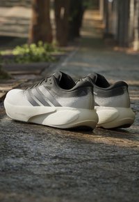 Paire de baskets athlétiques grises et blanches avec des semelles épaisses sur un pavement en pierre dans un cadre extérieur ombragé.
