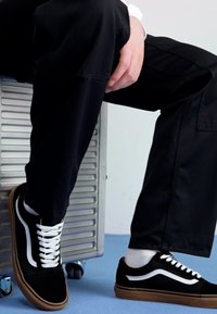 Svarte semskede sneakers med hvite striper, med snøring, gummisåle og teksturert stoff. Vist brukt med svarte bukser.