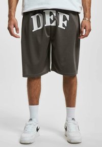 Grå treningsshorts med hvit påskrift "DEF" på framsiden, laget av et lett materiale. Kombinert med hvite sneakers og sokker.