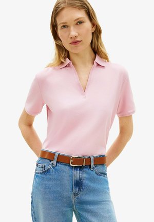 Femme portant un polo rose clair à manches courtes, rentré dans un jean bleu avec une ceinture marron, debout les mains dans le dos.