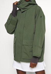 Veste verte imperméable avec col haut, coupe surdimensionnée, grandes poches, détails en velcro sur les manches et surface en tissu texturé.