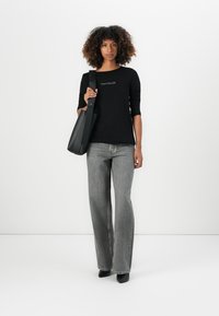 Black long-sleeve top with logo, gray flared jeans, and pointed black heels. Models a black shoulder bag, set against a minimal white background.