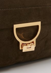 Zamszowa kopertówka w ciemnozielonym kolorze z złotym metalowym zapięciem z okrągłym wycięciem i wygrawerowanym logo. Teksturowana powierzchnia z kontrastującym przeszyciem.