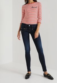 Woman wearing a red and white striped long-sleeve top with "Mademoiselle" text, dark skinny jeans, and black slip-on loafers.