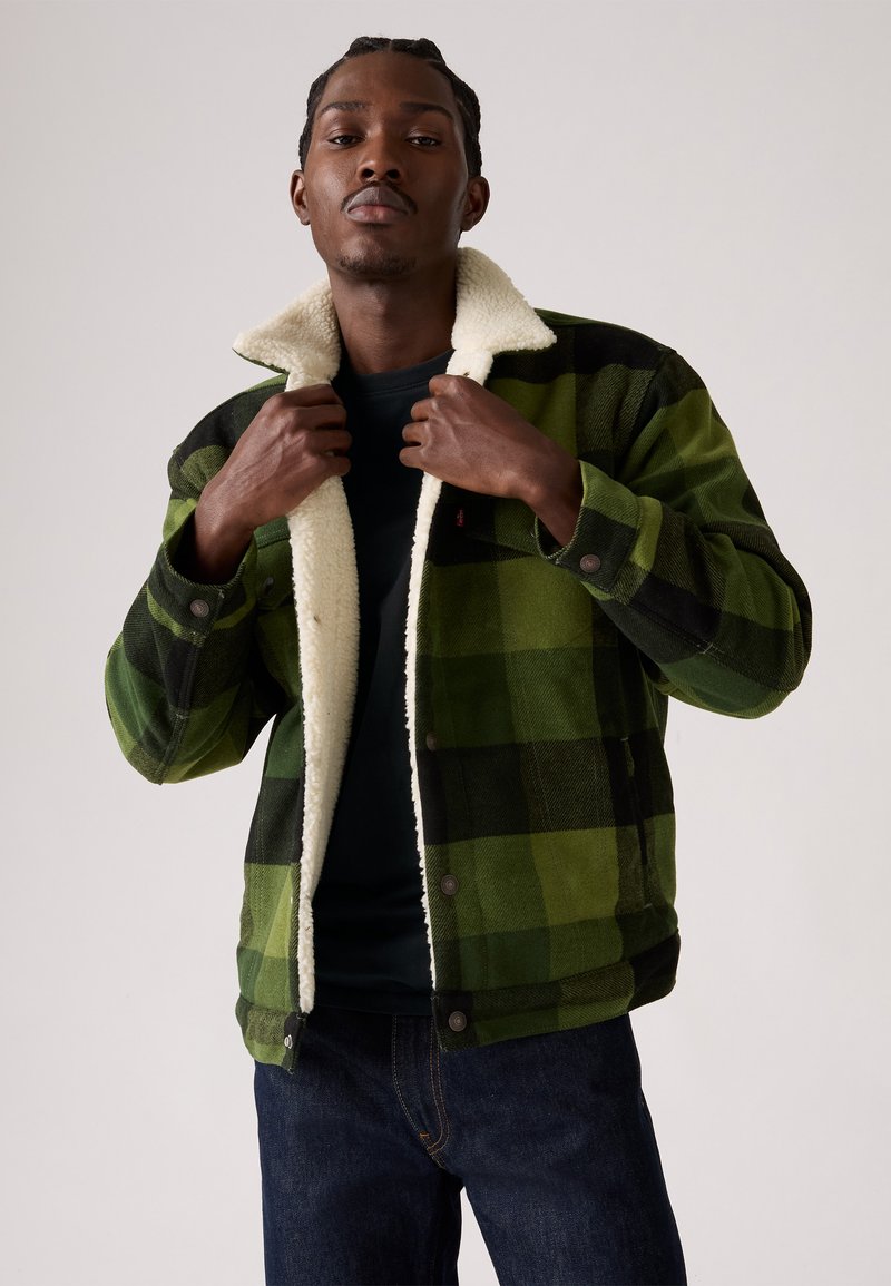 Groen en zwart geblokt flanellen jack met een beige sherpa voering, met drukknopen en lange mouwen, gedragen door een model.