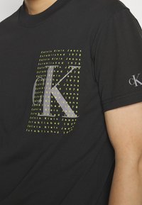 Black cotton t-shirt featuring a large graphic with "Calvin Klein Jeans" text in neon yellow and a stylized grey "K" on the front.