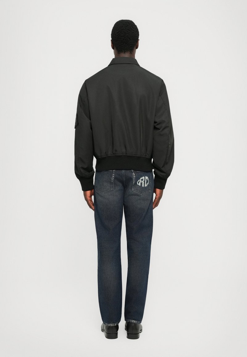 Blouson bomber noir avec un ourlet côtelé, porté avec un jean bleu foncé comportant des poches arrière et un léger délavage. Vue de dos.