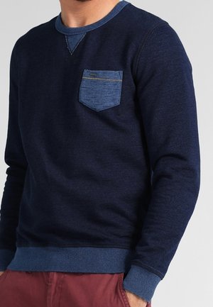 Man draagt een marineblauwe trui met lange mouwen, een lichterblauw borstzakje en geribbelde manchetten, gecombineerd met rode broek.