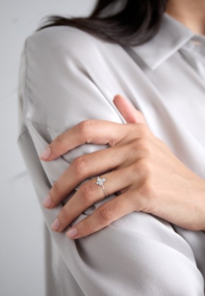 Anillo de plata con un diseño floral y pequeñas piedras preciosas, montado en una delicada cadena, llevado en una mano sobre una tela de color gris claro.