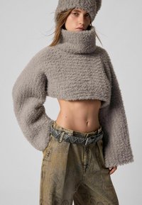 Pulover gri scurt cu textură pufoasă și guler înalt, asortat cu pantaloni din denim cu imprimeu, cu talie înaltă, și o centură decorativă.