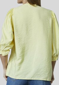 Gul bluse med en krøllet tekstur, 3/4 ærmer og rynkede detaljer ved manchetterne, stylet med blå jeans.