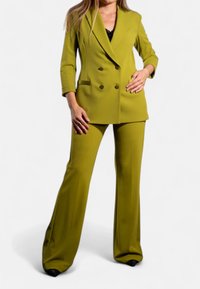 Abito doppiopetto in verde oliva, caratterizzato da un blazer su misura con bavero a punta e pantaloni a gamba larga. Tessuto liscio e leggero.