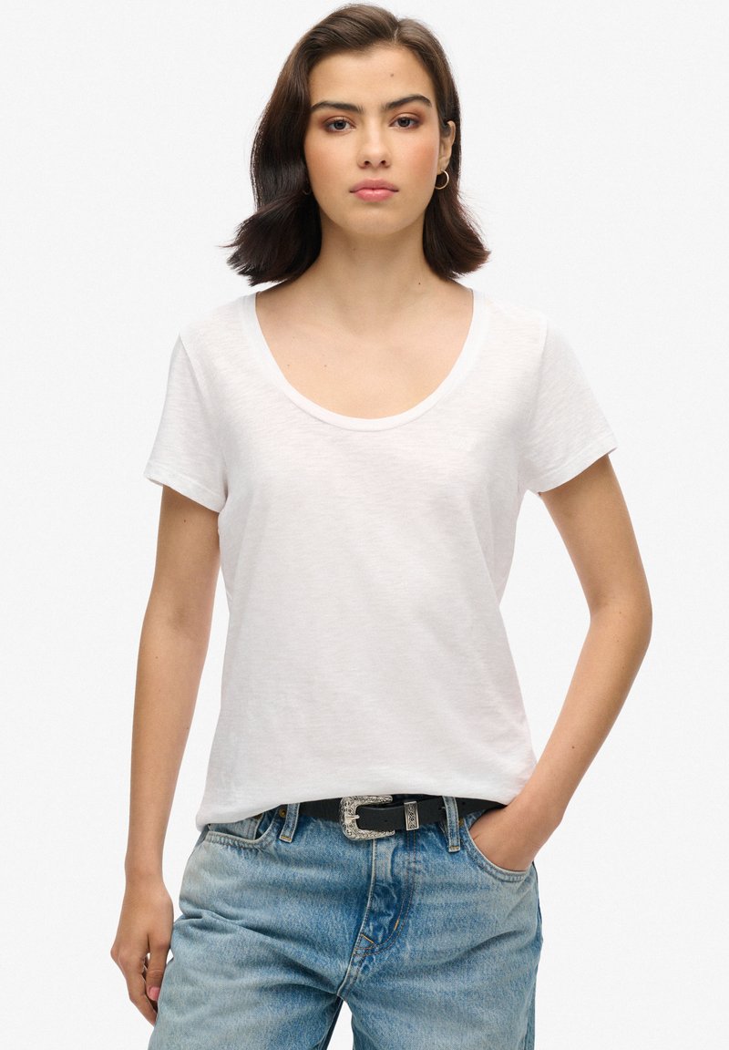 Camiseta blanca de escote redondo en tejido ligero con mangas cortas, combinada con jeans de mezclilla azules y un cinturón negro con hebilla plateada.