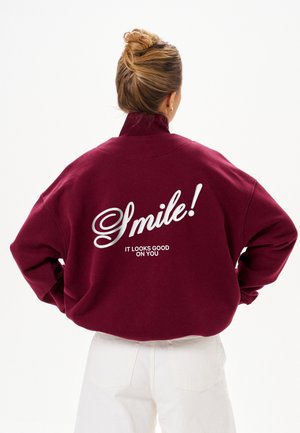 Femme aux cheveux blonds en chignon portant une veste bordeaux avec "Smile! It looks good on you" écrit dans le dos, associée à un pantalon blanc.